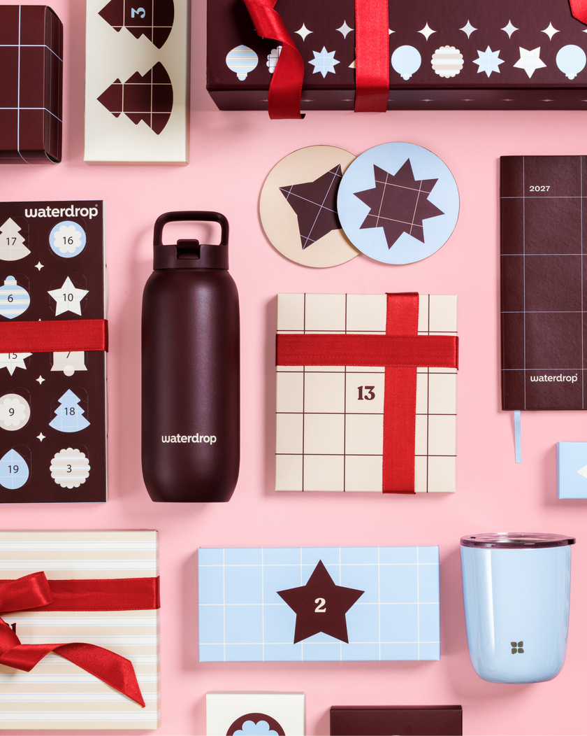 Assorted Advent calendars and gift boxes with red ribbons on a pink background, with a black water bottle and a light blue mug.