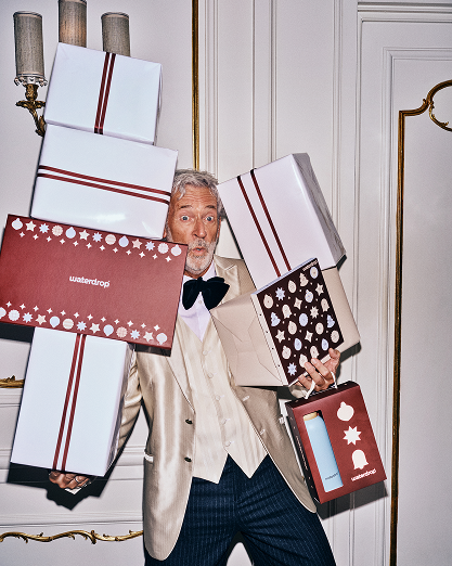 Man in a beige suit carries several wrapped gift boxes indoors.