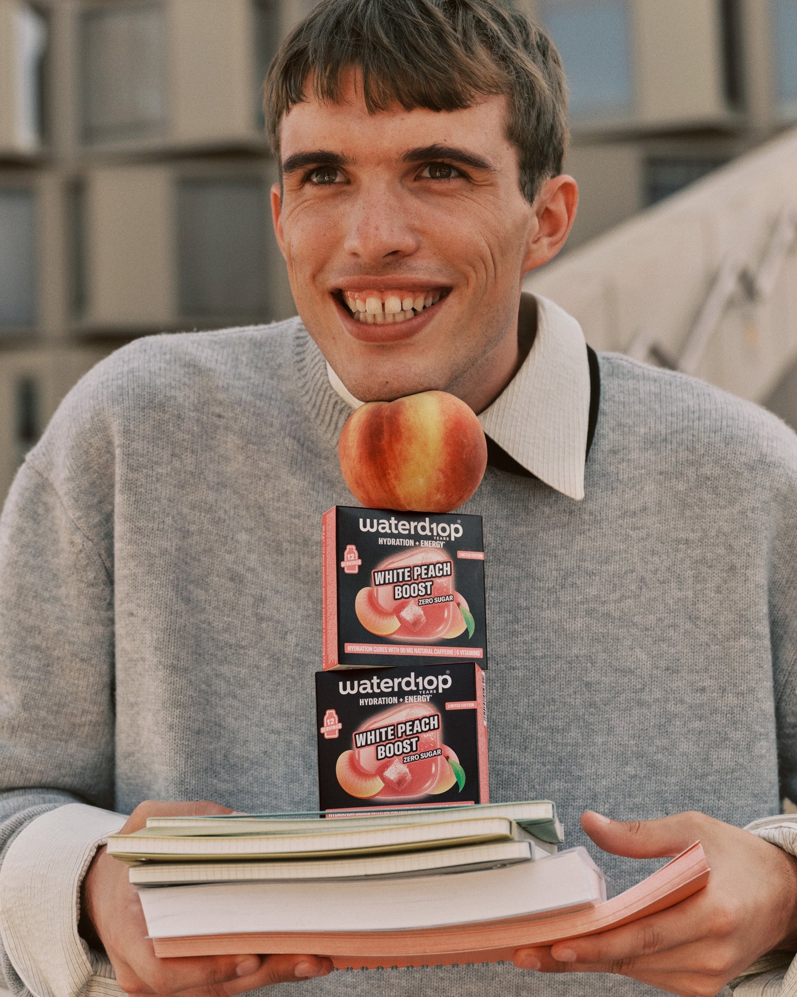 Young man in a gray jumper holding two peach-flavored boxes on top of a stack of books.
