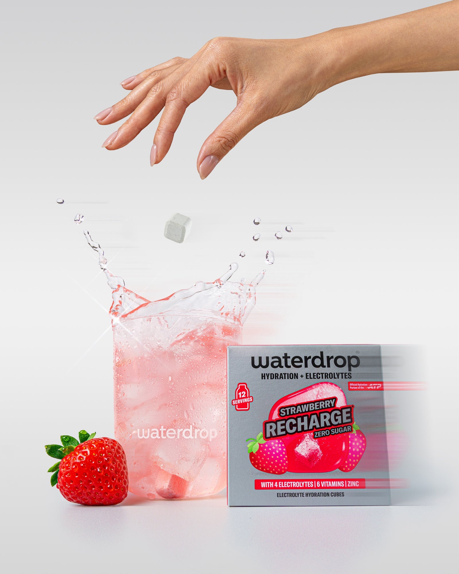 Hand dropping a white cube into a pink beverage beside a strawberry and a Waterdrop Strawberry Recharge box.
